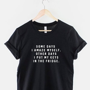 Può includere: T-shirt nero con la scritta "SOME DAYS I AMAZE MYSELF. OTHER DAYS I PUT MY KEYS IN THE FRIDGE." La maglietta è appesa a una gruccia di legno sospesa a una catena di metallo.