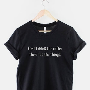May include: Black t-shirt with white text that reads "First I drink the coffee then I do the things."