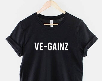 May include: Black t-shirt with the white text "VE-GAINZ" displayed prominently on the front. The shirt is hanging on a wooden hanger suspended from a metal chain. The sleeves are cuffed.