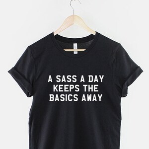 May include: Black t-shirt with white text that reads "A sass a day keeps the basics away".