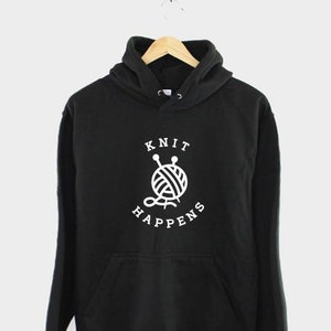 K&ouml;nnte beinhalten: Schwarzer Hoodie mit einem wei&szlig;en Aufdruck eines Wollkn&auml;uels und Stricknadeln mit dem Text "Knit Happens".