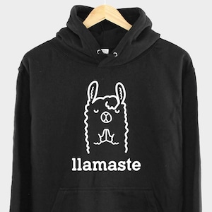 Op de afbeelding: Zwart hoodie met een witte omtrek van een mediterende lama en de tekst "llamaSTE" eronder.