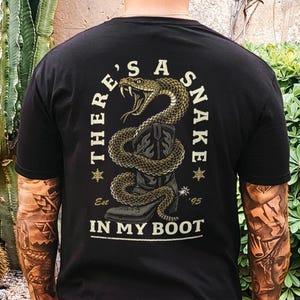 May include: Black t-shirt with a graphic of a coiled snake above a cowboy boot. The text "THERE'S A SNAKE IN MY BOOT" is arched above and below the snake design. The shirt is worn by a person in khaki pants.