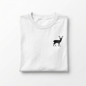 Puede incluir: Una camiseta blanca de cuello redondo que presenta una peque&ntilde;a silueta negra s&oacute;lida de un ciervo de pie con astas, bordada en la parte superior izquierda del pecho. La prenda est&aacute; cuidadosamente doblada, mostrando un dise&ntilde;o limpio.