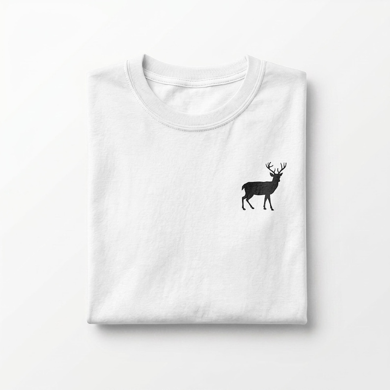 Puede incluir: Una camiseta blanca de cuello redondo que presenta una peque&ntilde;a silueta negra s&oacute;lida de un ciervo de pie con astas, bordada en la parte superior izquierda del pecho. La prenda est&aacute; cuidadosamente doblada, mostrando un dise&ntilde;o limpio.