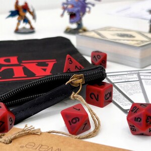 May include: A black fabric pouch with a red logo and a brass zip pull, accompanied by several red polyhedral dice with black numbers. The dice include a d4, d6, d8, and d20, scattered alongside a deck of cards and game miniatures.