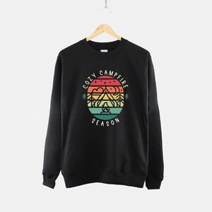 Op de afbeelding: Zwart sweatshirt met een afbeelding van een berglandschap met een kampvuur en de tekst "Cozy Campfire Season" in een cirkel.