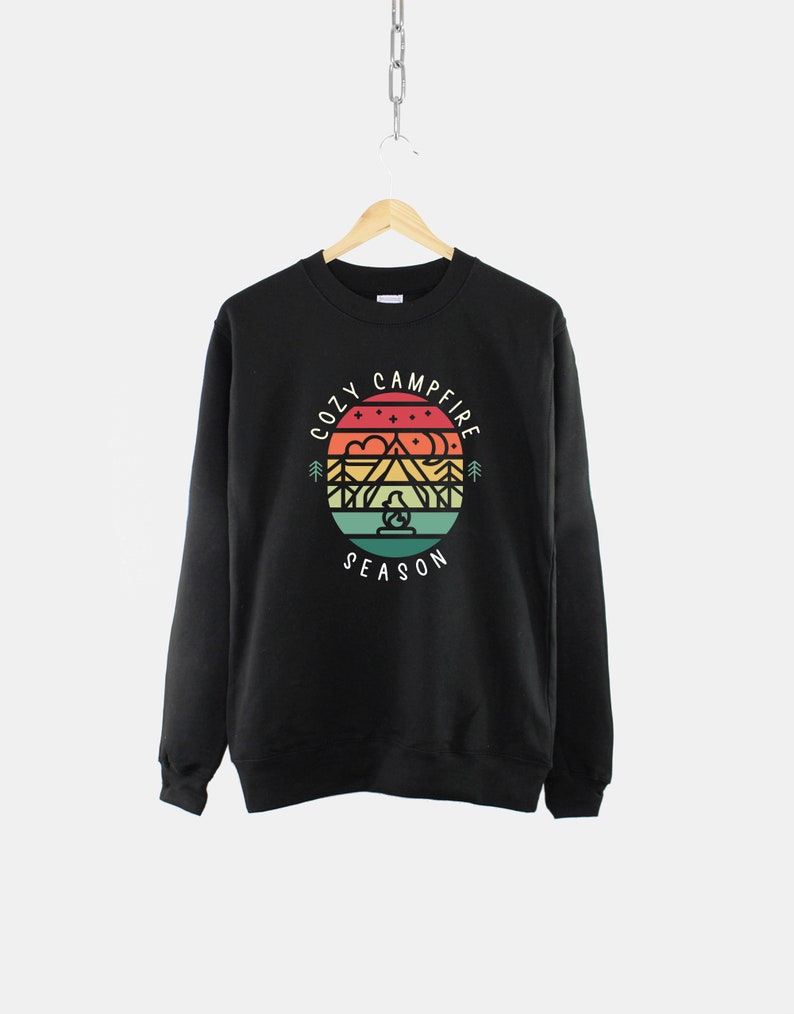 Op de afbeelding: Zwart sweatshirt met een afbeelding van een berglandschap met een kampvuur en de tekst "Cozy Campfire Season" in een cirkel.