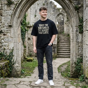 May include: A black short-sleeved t-shirt featuring the white text "WHEN THE DM SMILES, IT'S ALREADY TOO LATE" printed on the chest, paired with dark gray denim jeans and white sneakers.