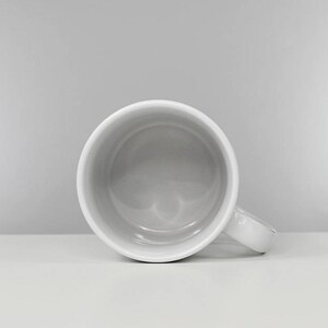 May include: A white ceramic mug with a rounded handle. The mug is empty and has a smooth, glossy finish.