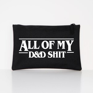 May include: A black rectangular zippered pouch featuring white text that reads ALL OF MY D&D SHIT. The text is centred on the front of the pouch, with horizontal lines above and below the words ALL OF MY, creating a bold graphic design.