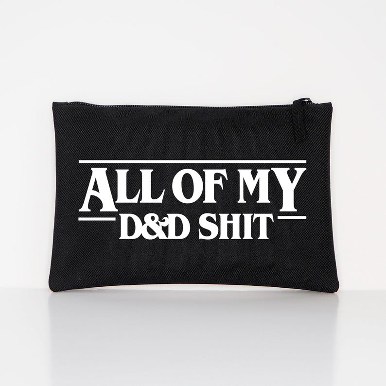 May include: A black rectangular zippered pouch featuring white text that reads ALL OF MY D&D SHIT. The text is centred on the front of the pouch, with horizontal lines above and below the words ALL OF MY, creating a bold graphic design.