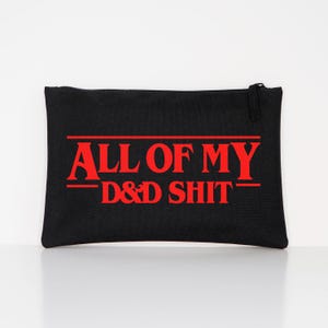 May include: A black rectangular zippered pouch featuring the red text ALL OF MY D&D SHIT printed on the front. The text is arranged in three lines with horizontal red lines above and below the words, creating a bold graphic design on the fabric.