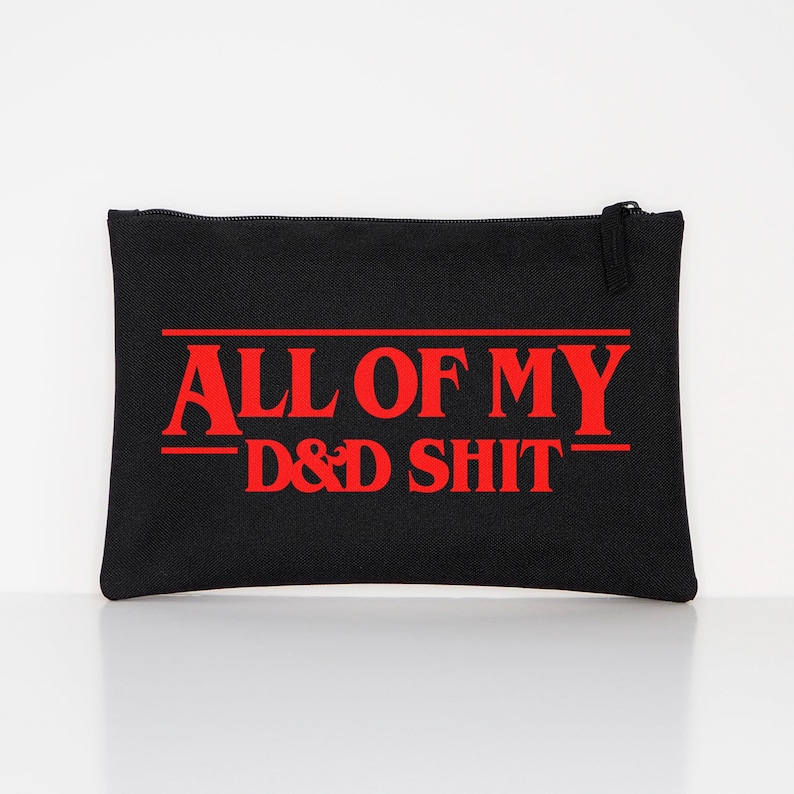 May include: A black rectangular zippered pouch featuring the red text ALL OF MY D&D SHIT printed on the front. The text is arranged in three lines with horizontal red lines above and below the words, creating a bold graphic design on the fabric.