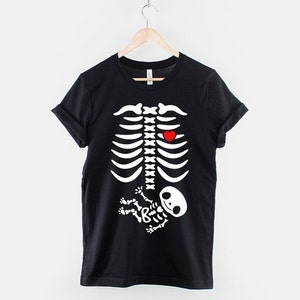 May include: Black t-shirt with a white skeleton graphic. The skeleton has a red heart in its chest and a smaller white skeleton baby in its belly.