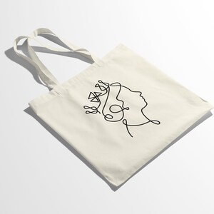 Op de afbeelding: Een witte tote bag met een zwarte lijntekening van een gezicht met een kroon op het hoofd.