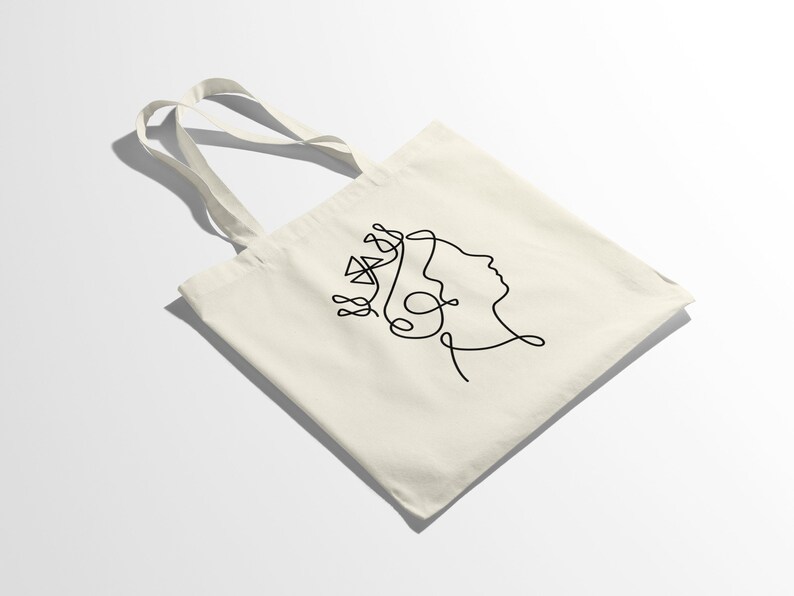 Op de afbeelding: Een witte tote bag met een zwarte lijntekening van een gezicht met een kroon op het hoofd.