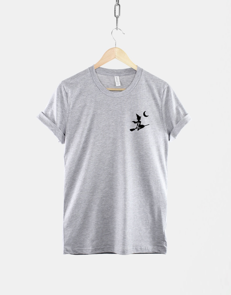 Op de afbeelding: Een heidekleurig t-shirt met een zwart silhouet van een heks die op een bezem vliegt met een halve maan op de achtergrond.