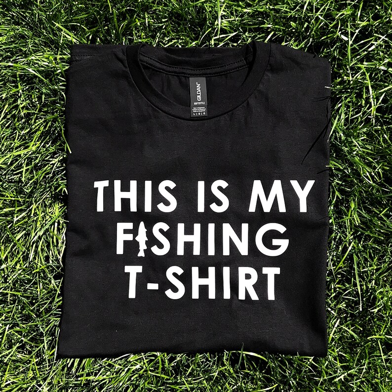 Może przedstawiać: Czarny T-shirt z białym napisem "This is my fishing t-shirt" i stylizowanym wzorem ryby.