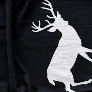 Puede incluir: Una prenda de tela negra que presenta una gran silueta blanca serigrafiada de un ciervo con astas prominentes. El ciervo se muestra en una pose din&aacute;mica y saltarina, con la cabeza vuelta hacia arriba sobre la superficie textil oscura.