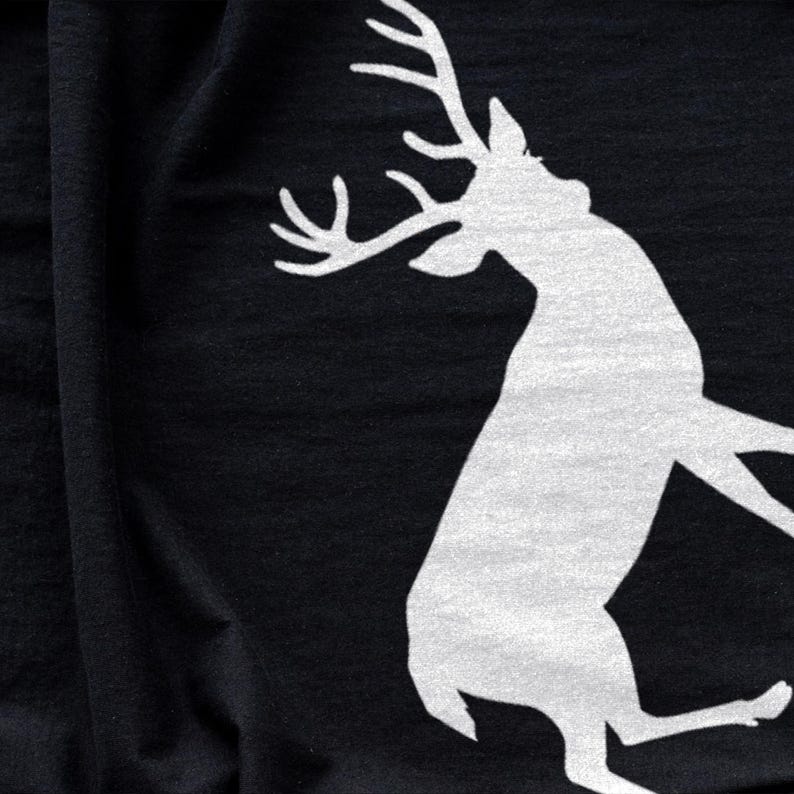 Puede incluir: Una prenda de tela negra que presenta una gran silueta blanca serigrafiada de un ciervo con astas prominentes. El ciervo se muestra en una pose din&aacute;mica y saltarina, con la cabeza vuelta hacia arriba sobre la superficie textil oscura.