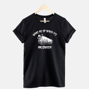 Puede incluir: Camiseta negra con un gr&aacute;fico blanco de un esqueleto en un ata&uacute;d. El texto "Wake me up when it's Halloween" est&aacute; impreso alrededor del gr&aacute;fico.