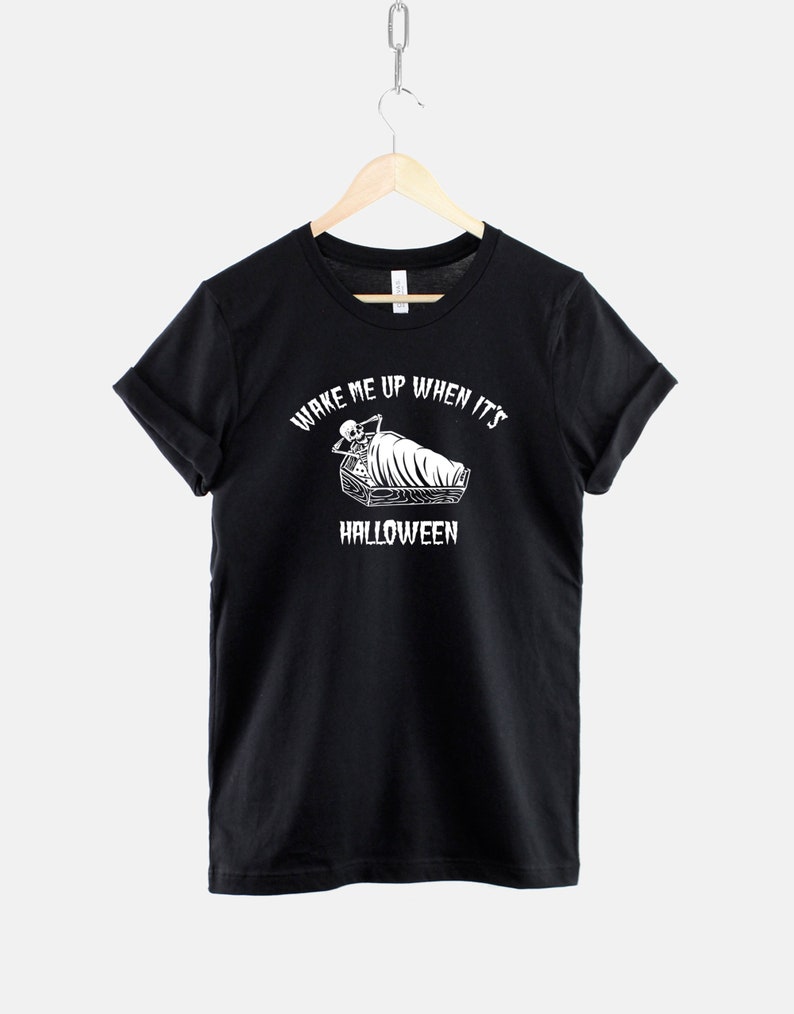 Puede incluir: Camiseta negra con un gr&aacute;fico blanco de un esqueleto en un ata&uacute;d. El texto "Wake me up when it's Halloween" est&aacute; impreso alrededor del gr&aacute;fico.