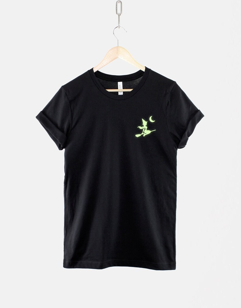 Op de afbeelding: Zwart T-shirt met een groene heks silhouette op een bezem die langs een halve maan vliegt.