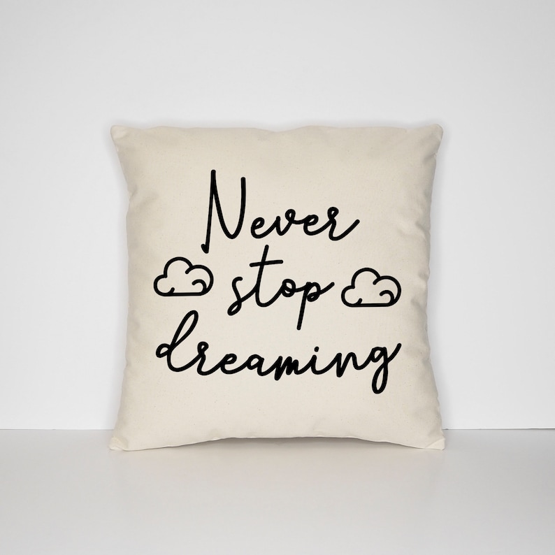 May include: A white throw pillow with the text "Never stop dreaming" in black cursive font. Two small cloud drawings are on either side of the text.
