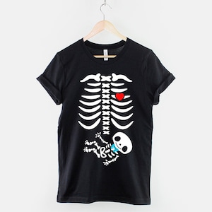 May include: Black t-shirt with a white skeleton graphic. The skeleton has a red heart and a small baby skeleton with a blue bow tie inside its ribcage.