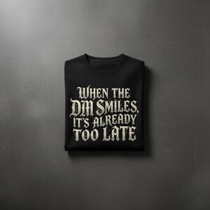 May include: A black short-sleeved t-shirt featuring white gothic-style lettering that reads, "WHEN THE DM SMILES, IT'S ALREADY TOO LATE." The text is centered on the chest of the folded garment, which has a classic crew neckline and a smooth fabric finish.