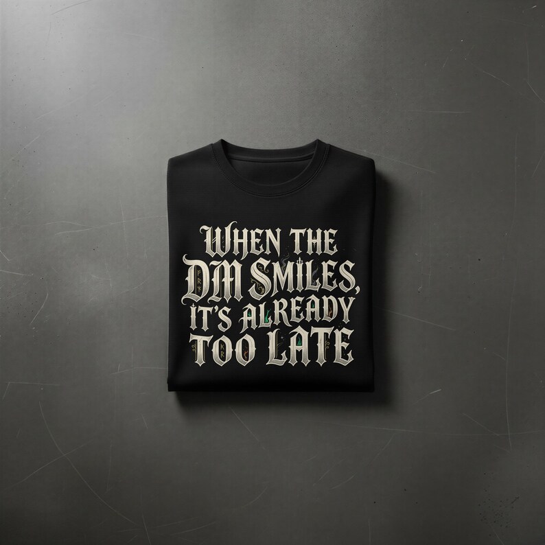 May include: A black short-sleeved t-shirt featuring white gothic-style lettering that reads, "WHEN THE DM SMILES, IT'S ALREADY TOO LATE." The text is centered on the chest of the folded garment, which has a classic crew neckline and a smooth fabric finish.