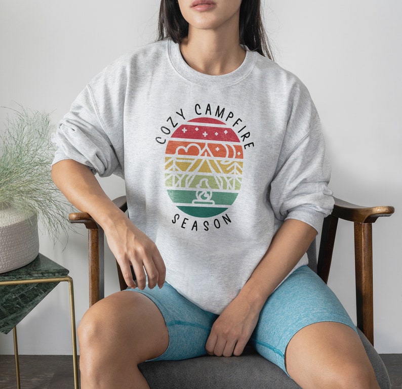 Op de afbeelding: Een grijs sweatshirt met een retro-grafisch ontwerp met een kampvuursc&egrave;ne en de tekst "Cozy Campfire Season".