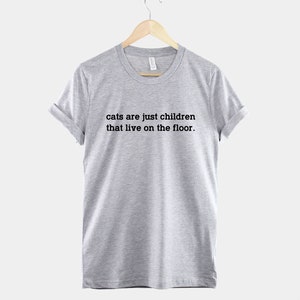 May include: A heather grey t-shirt with the text "cats are just children that live on the floor." printed in black.