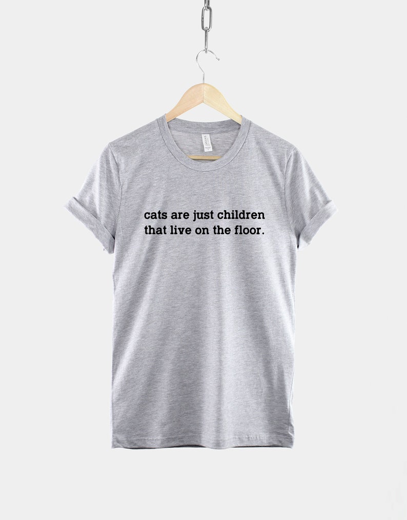 May include: A heather grey t-shirt with the text "cats are just children that live on the floor." printed in black.