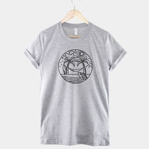 May include: A heather grey t-shirt with a black and white illustration of palm trees, a hammock, a surfboard, and a sun. The illustration is inside a circle.