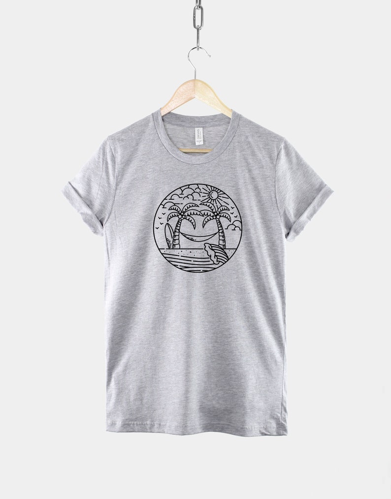 May include: A heather grey t-shirt with a black and white illustration of palm trees, a hammock, a surfboard, and a sun. The illustration is inside a circle.