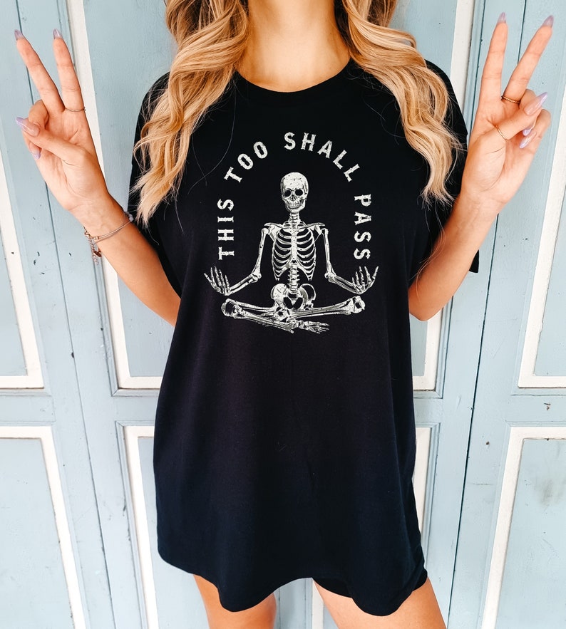 May include: Black t-shirt with a white graphic of a skeleton sitting in a yoga pose with the text "This Too Shall Pass" around it.