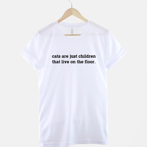 May include: White t-shirt with black text that reads "cats are just children that live on the floor."
