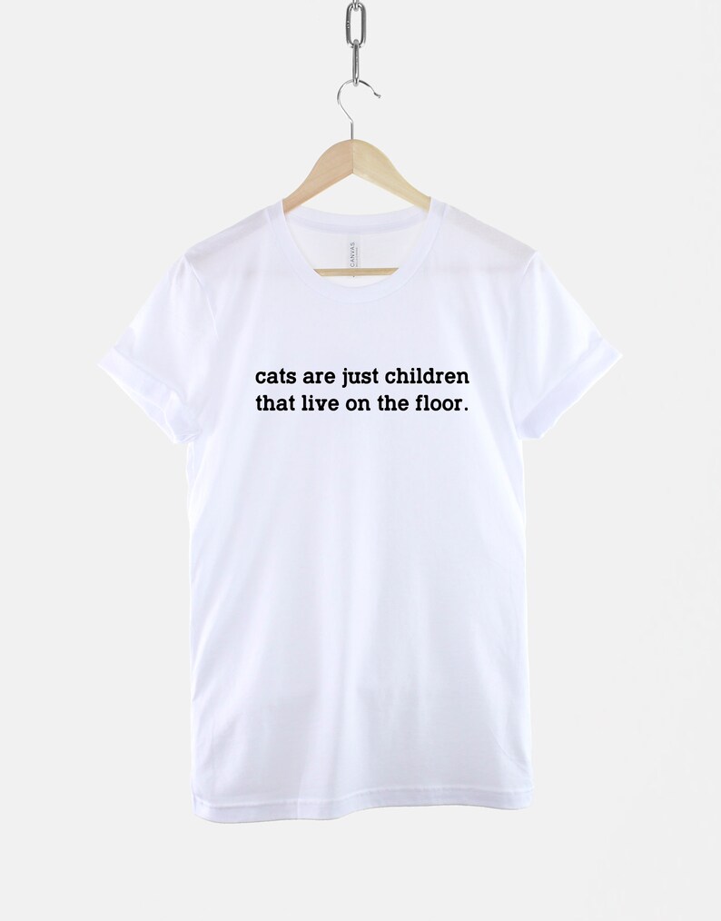 May include: White t-shirt with black text that reads "cats are just children that live on the floor."