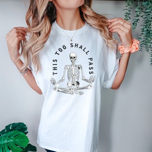 May include: White t-shirt with a black skeleton meditating in a yoga pose. The text "This too shall pass" is written in a circle around the skeleton.
