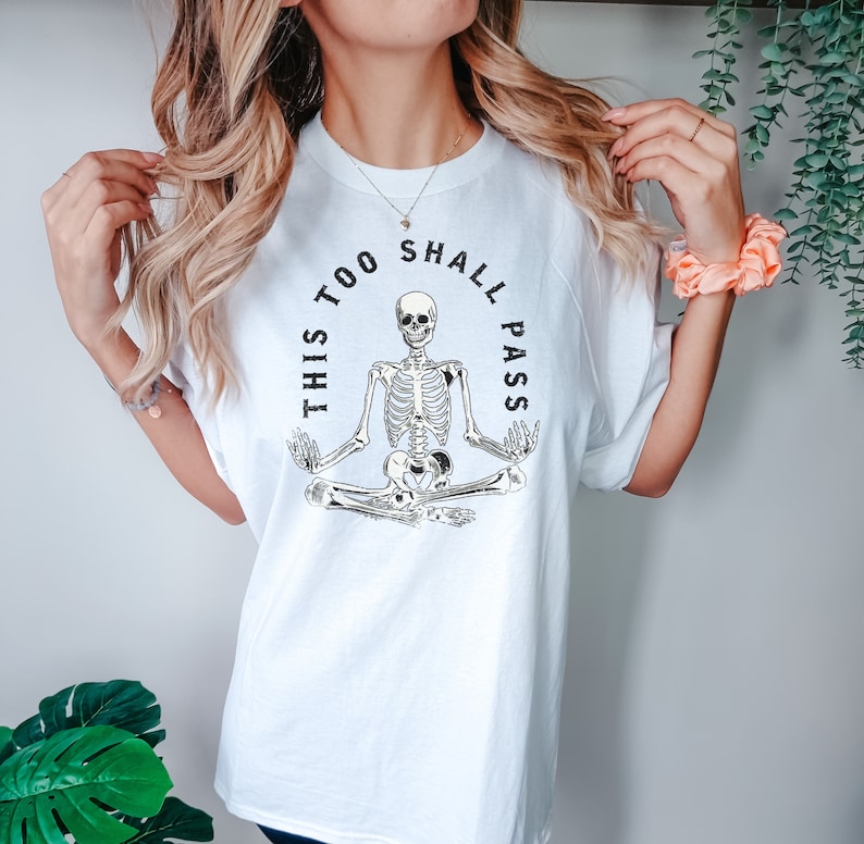 May include: White t-shirt with a black skeleton meditating in a yoga pose. The text "This too shall pass" is written in a circle around the skeleton.