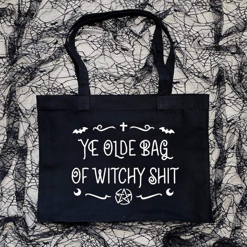 Puede incluir: Bolsa de tela negra con texto blanco que dice "Ye Olde Bag of Witchy Shit". El texto est&aacute; rodeado por un borde decorativo con murci&eacute;lagos, una cruz y un pentagrama.