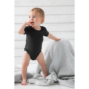 May include: A baby wearing a black short-sleeved bodysuit stands on a gray blanket. The baby is looking to the right of the frame.