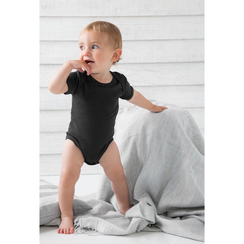 May include: A baby wearing a black short-sleeved bodysuit stands on a gray blanket. The baby is looking to the right of the frame.