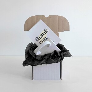 Puede incluir: Una taza de cer&aacute;mica blanca con asa est&aacute; anidada en papel de seda negro dentro de una caja de cart&oacute;n blanca. Una tarjeta blanca con el texto "thank you." es visible en la parte superior de la caja.