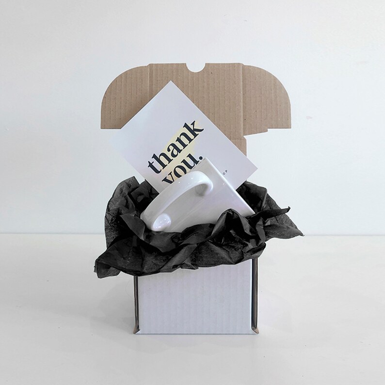 Puede incluir: Una taza de cer&aacute;mica blanca con asa est&aacute; anidada en papel de seda negro dentro de una caja de cart&oacute;n blanca. Una tarjeta blanca con el texto "thank you." es visible en la parte superior de la caja.