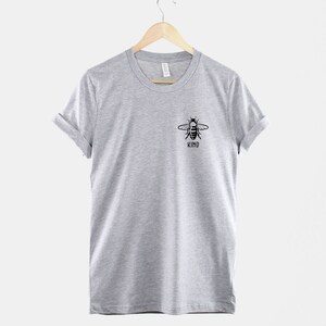 Op de afbeelding: Een grijs t-shirt met een zwarte bij-afbeelding en het woord "KIND" eronder.
