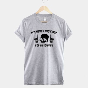 Peut inclure: T-shirt gris avec un motif de cr&acirc;ne et d'os crois&eacute;s noirs. Le texte "It's never too early for Halloween" est imprim&eacute; sous le cr&acirc;ne.