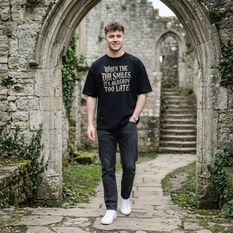 May include: A black short-sleeved t-shirt featuring the text "WHEN THE DM SMILES, IT'S ALREADY TOO LATE" in a distressed white font on the chest, paired with dark gray denim jeans and white sneakers.